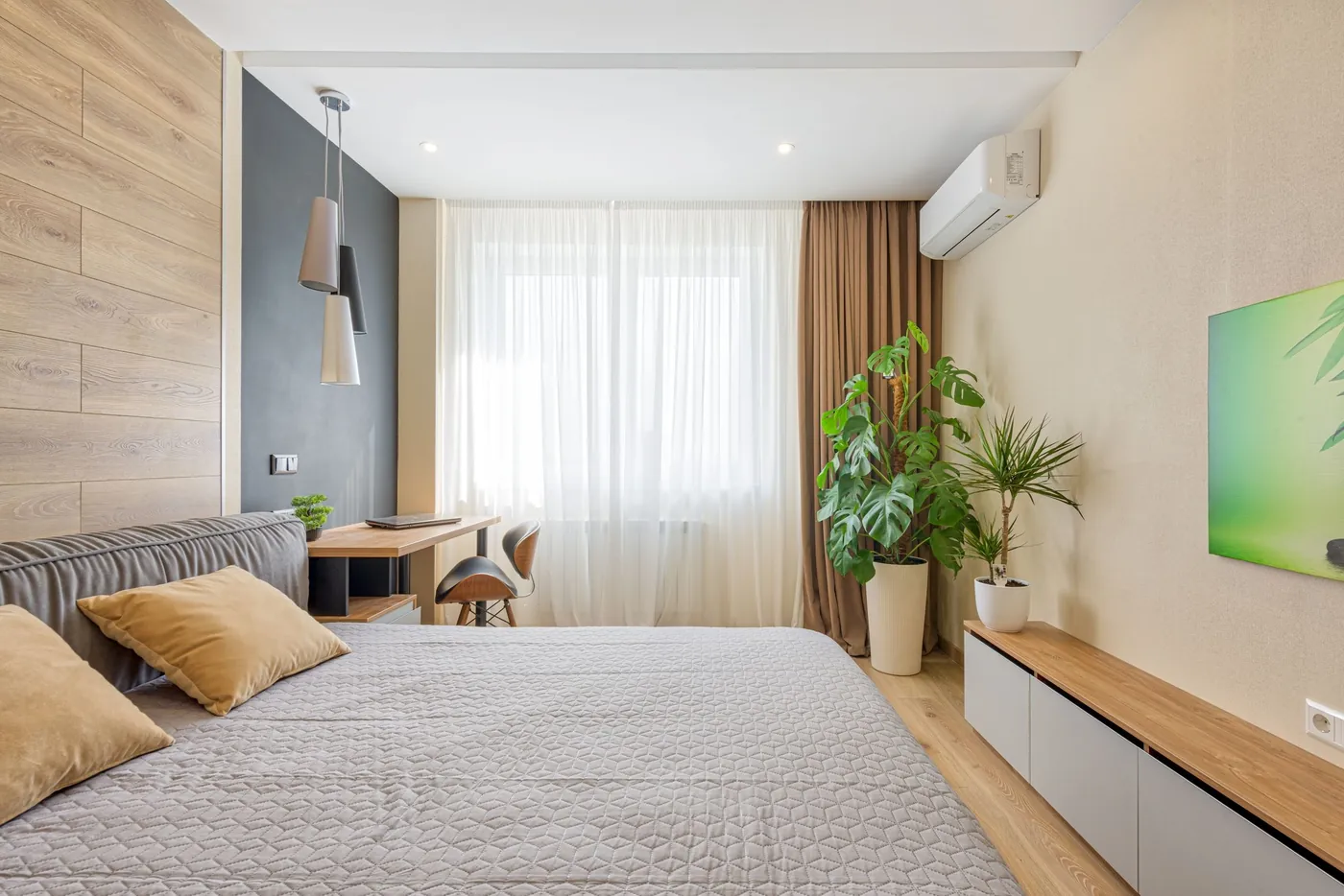 Chambre contemporaine aux tons sable et bois clair, lit gris, bureau, grandes plantes et unité intérieure murale près des voilages
