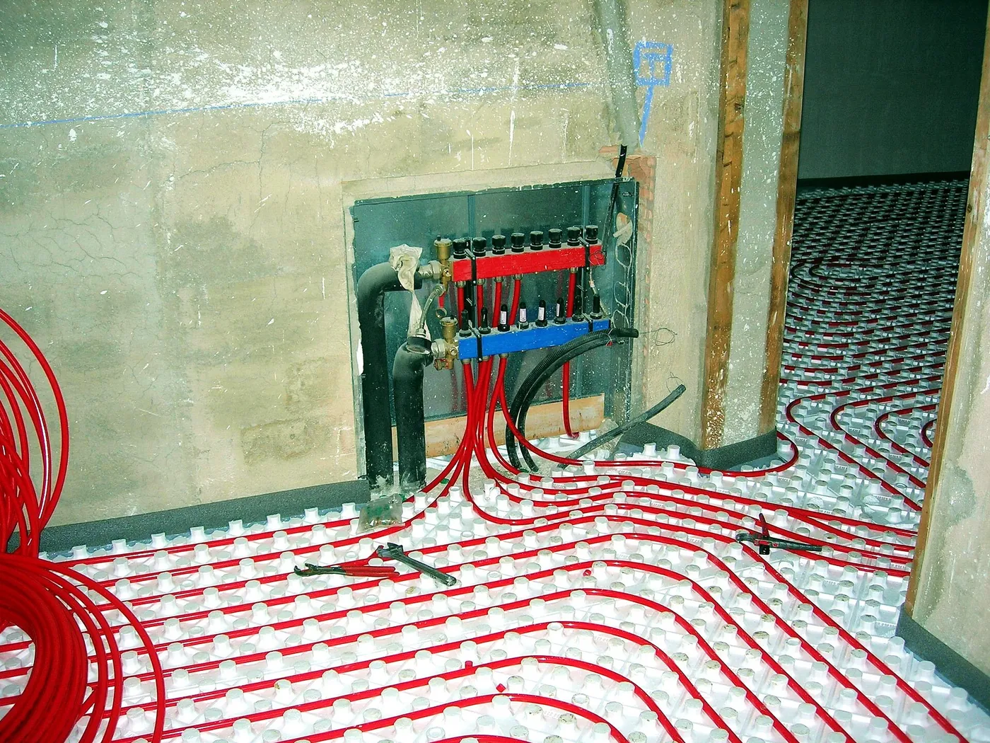Collecteur rouge et bleu dans une niche murale, d'où partent les tubes rouges du plancher chauffant posés sur plaques à plots