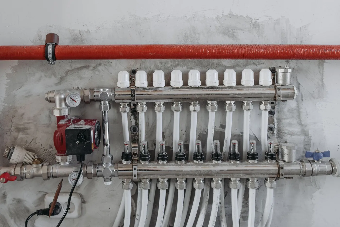 Collecteur inox de plancher chauffant équipé d'un circulateur rouge et d'une rangée de débitmètres, fixé sur un mur brut