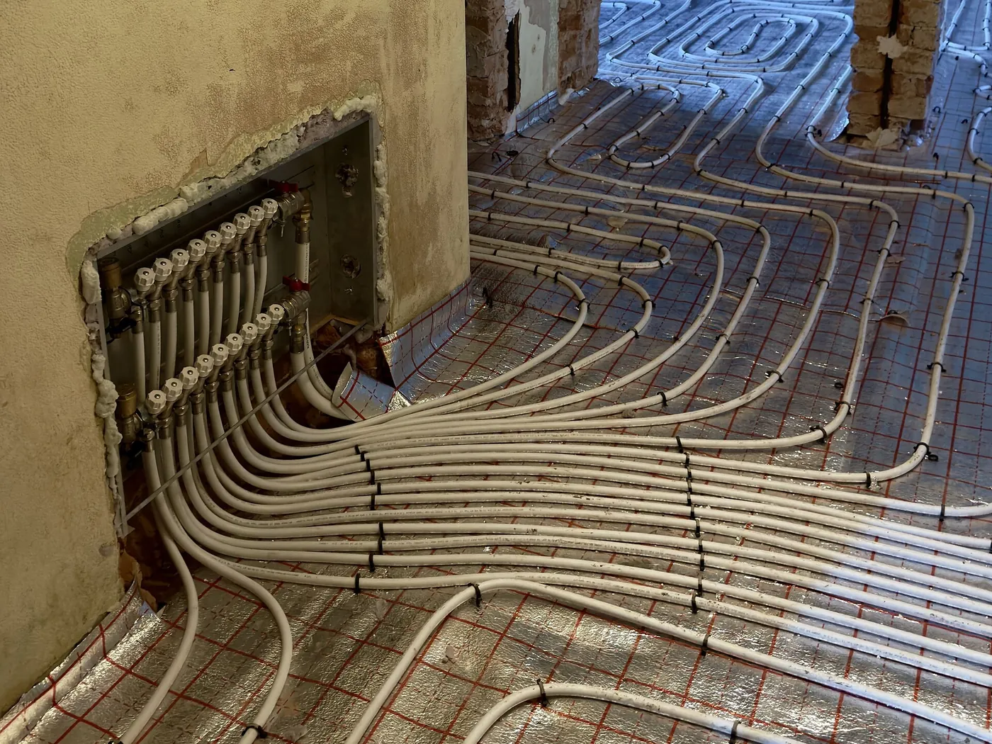 Collecteur encastré dans un mur ancien d'où rayonnent des dizaines de tubes blancs de plancher chauffant sur isolant aluminium