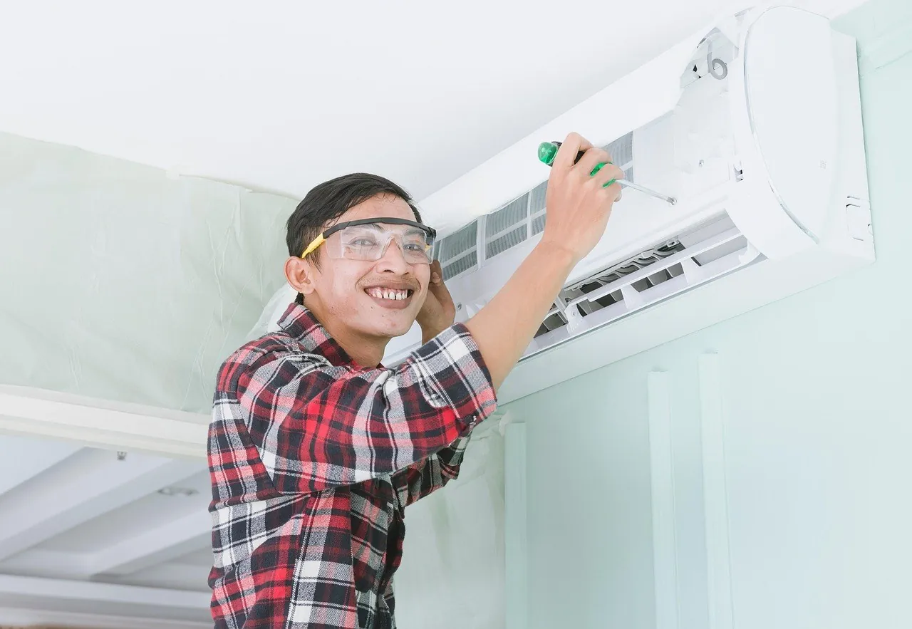 Installateur souriant en chemise à carreaux et lunettes de protection, réglant au tournevis une unité intérieure murale ouverte
