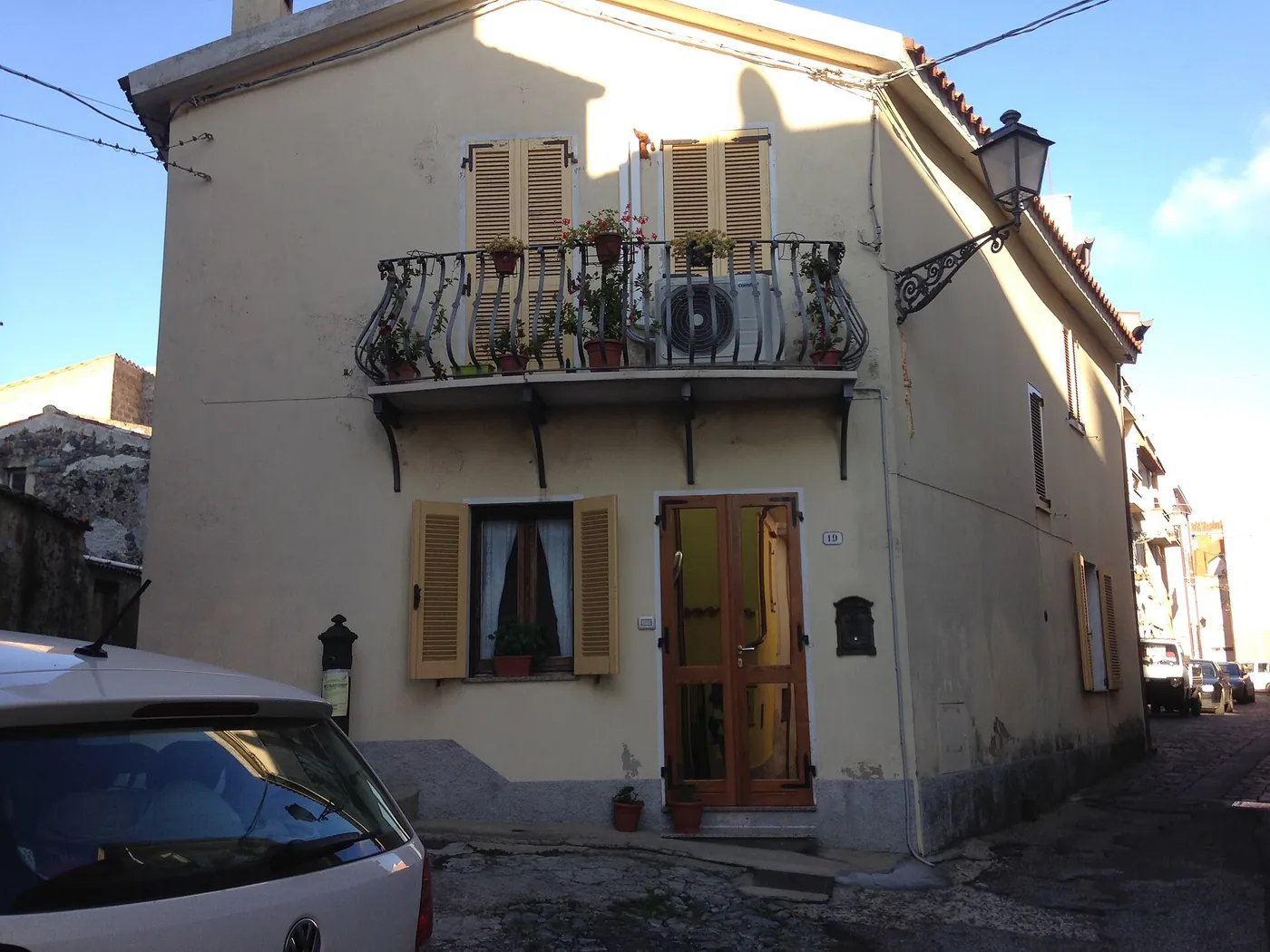 Maison de village crème aux volets en bois, unité extérieure posée sur un balcon en fer forgé fleuri, au bord d'une ruelle pavée