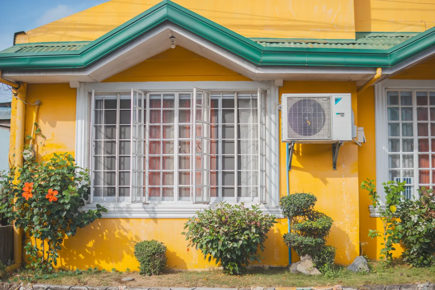 Maison au crépi jaune vif à grandes fenêtres blanches, unité extérieure sur consoles murales, hibiscus en fleurs et arbustes taillés