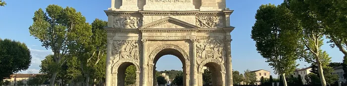 Arc de triomphe romain d'Orange en pierre claire sculptée, entouré de platanes, sous un ciel bleu d'été