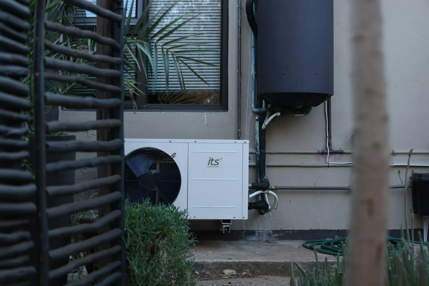 Pompe à chaleur monobloc blanche et son ballon noir fixé au mur, tuyauteries, fenêtre avec palmier, claustra en bois au premier plan