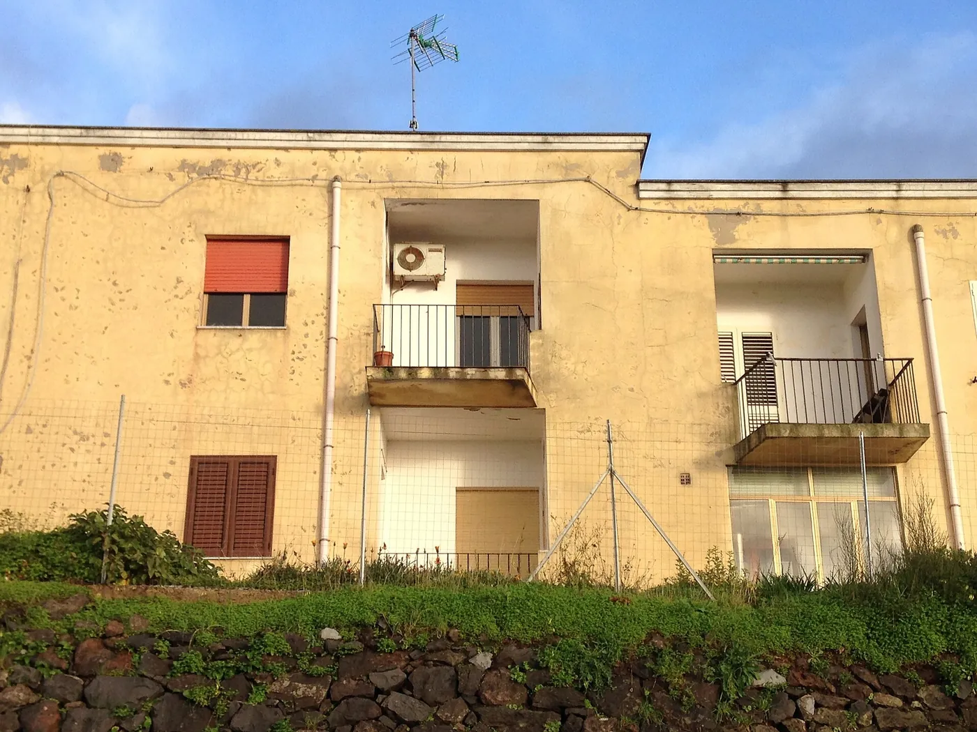 Petit immeuble à l'enduit ocre défraîchi, unité extérieure dans la loggia de l'étage, balcons à garde-corps, muret de pierres sèches