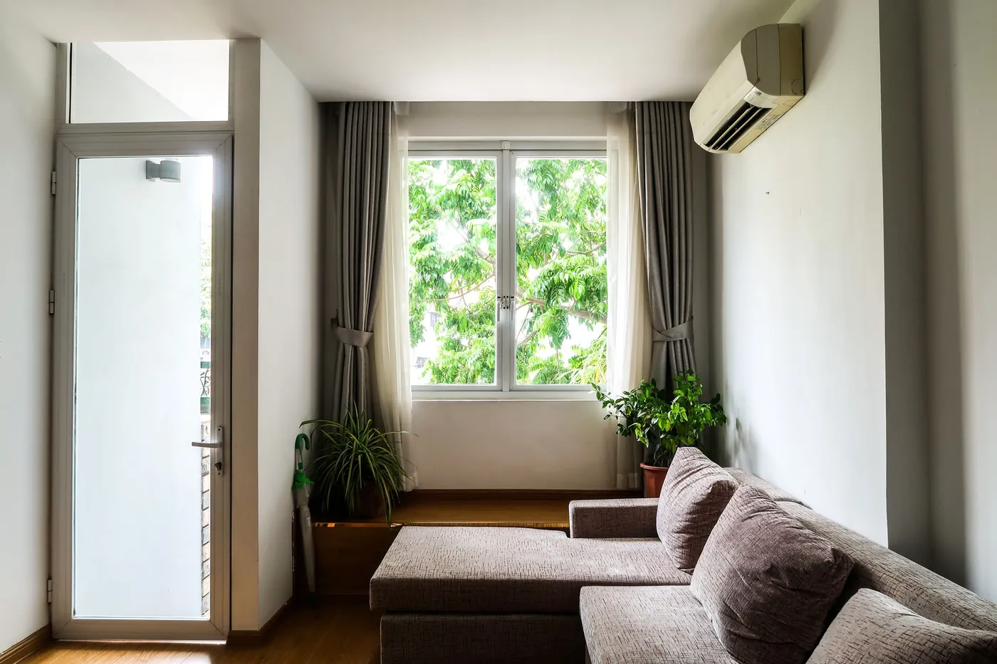 Petit salon clair avec canapé méridienne gris, plantes vertes, rideaux gris et unité intérieure murale près d'une fenêtre sur les arbres