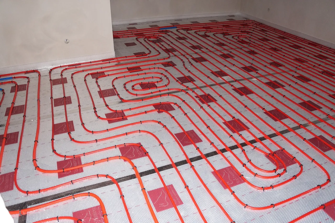 Tubes rouges de plancher chauffant disposés en spirale sur un isolant aluminium à repères rouges, dans une pièce vide