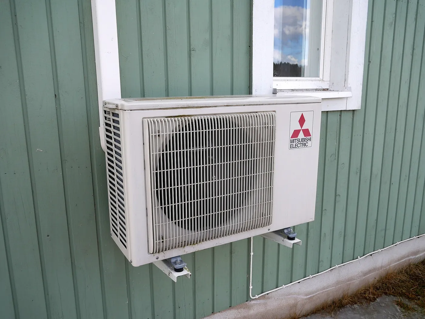 Unité extérieure de pompe à chaleur air/air sur consoles sous une fenêtre, contre une maison en bardage de bois peint en vert