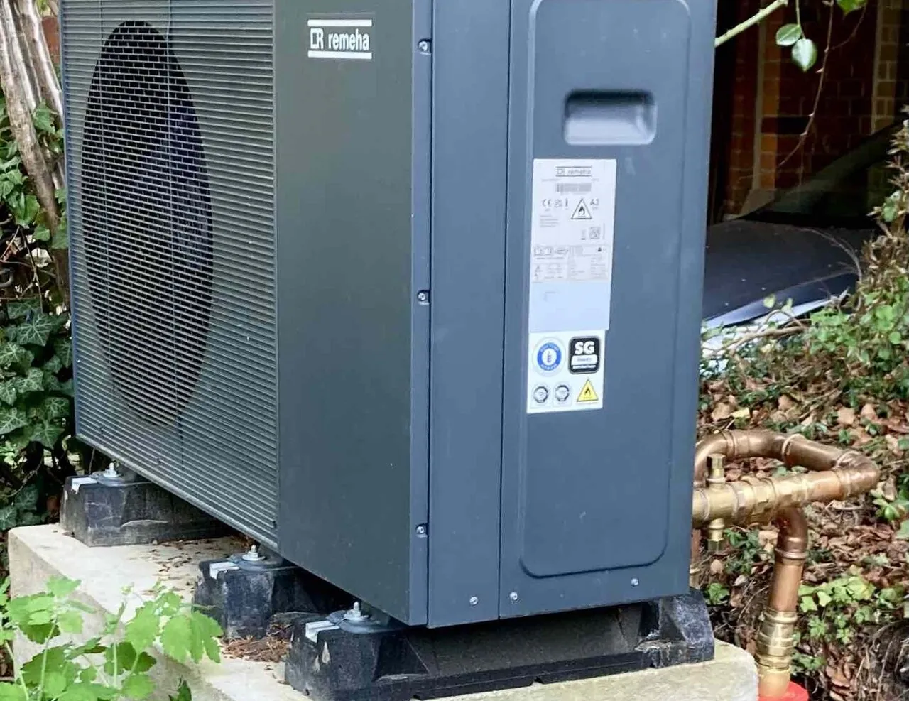 Unité extérieure anthracite de pompe à chaleur air/eau sur socle en béton, entourée de lierre, tuyaux en cuivre au premier plan