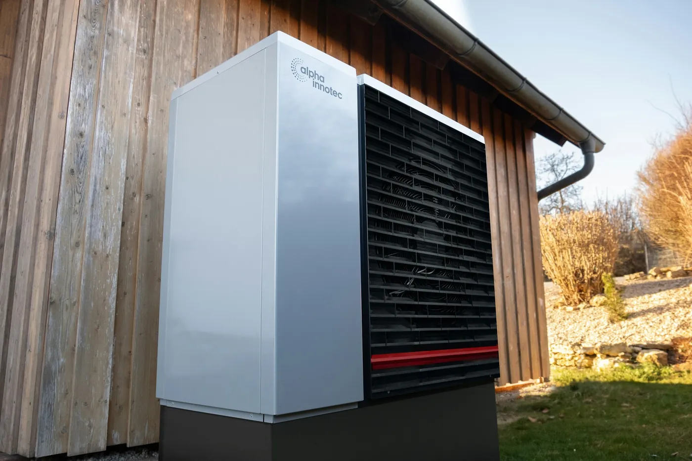 Unité extérieure blanche et noire de pompe à chaleur posée contre le bardage en bois d'une maison, sous un ciel bleu d'automne