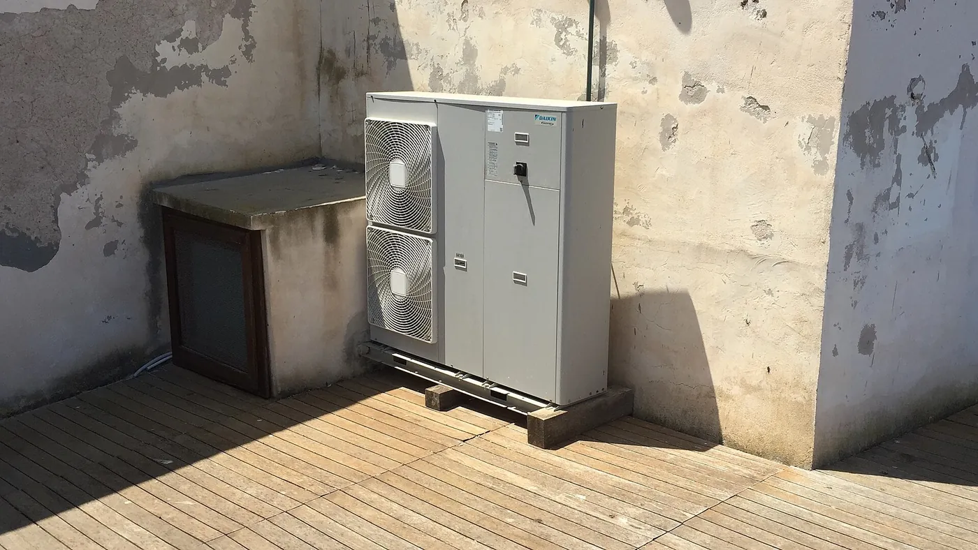Unité extérieure à double ventilateur posée sur cales en bois, sur un plancher de lattes, dans une cour aux vieux murs écaillés