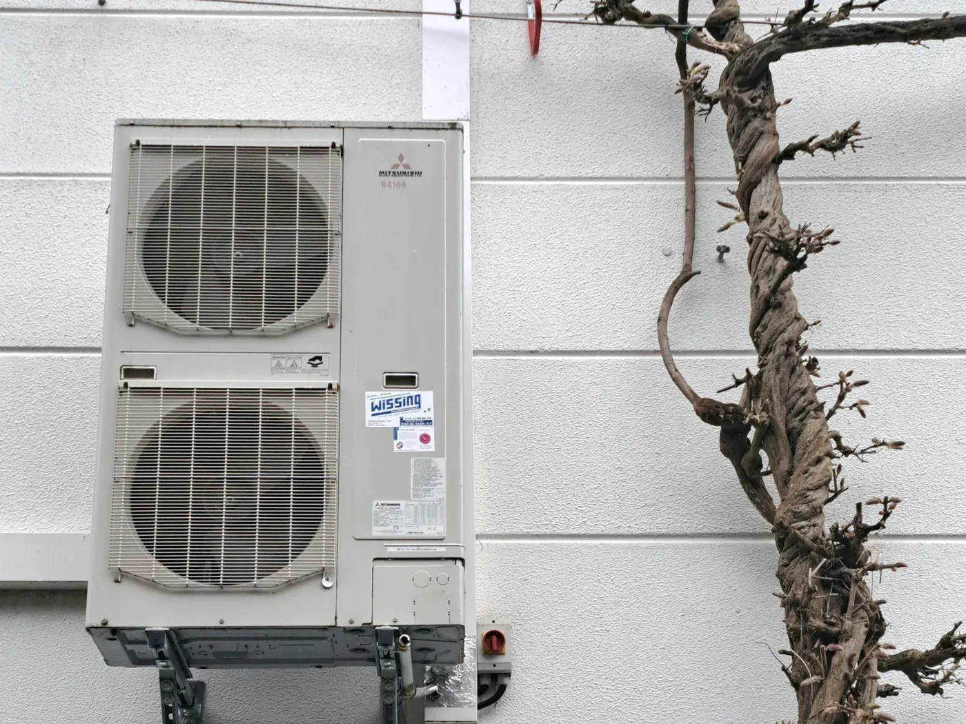 Unité extérieure à double ventilateur sur consoles contre une façade blanche, à côté du tronc noueux d'une glycine