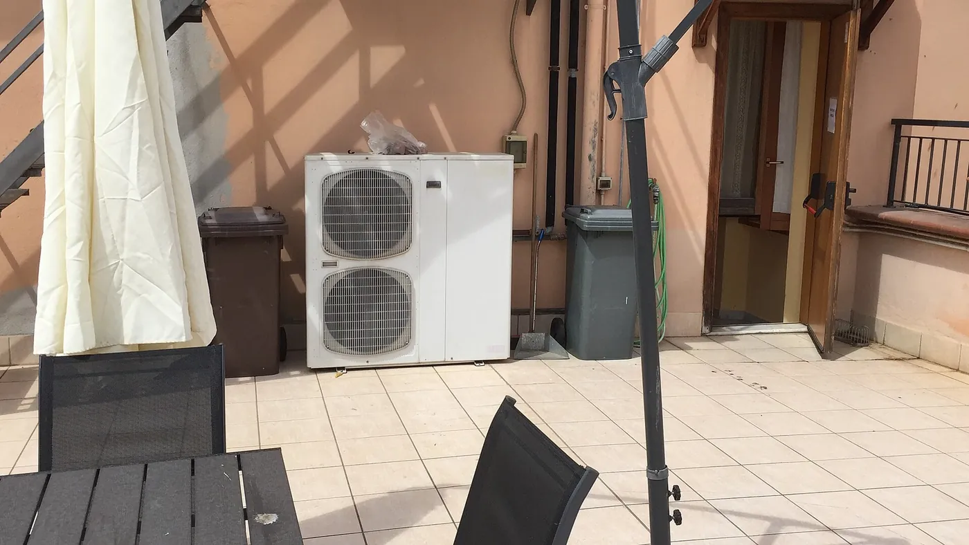 Unité extérieure de pompe à chaleur à deux ventilateurs posée sur une terrasse carrelée, près d'un salon de jardin