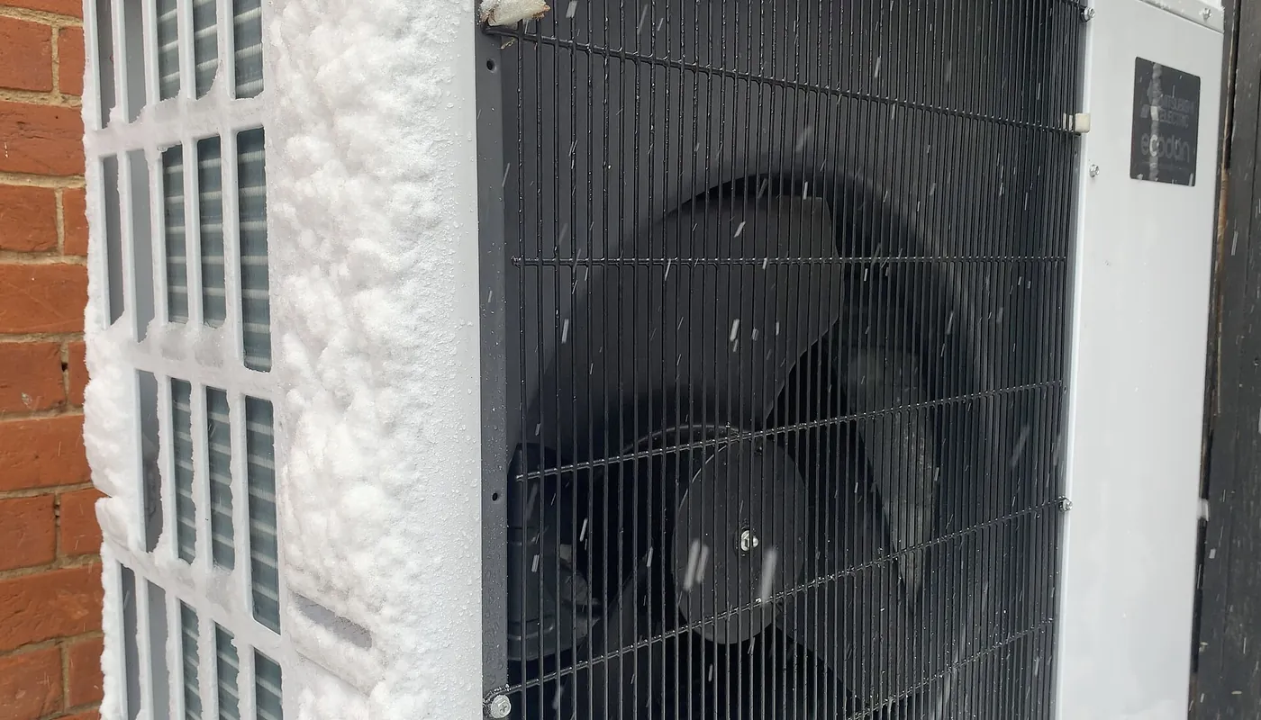 Unité extérieure de pompe à chaleur couverte de givre épais contre un mur de brique, pendant une chute de neige