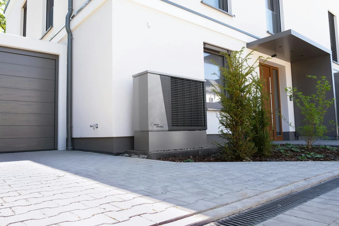 Pompe à chaleur air-eau grise posée au pied d'une maison neuve blanche, entre la porte de garage et l'entrée, allée pavée