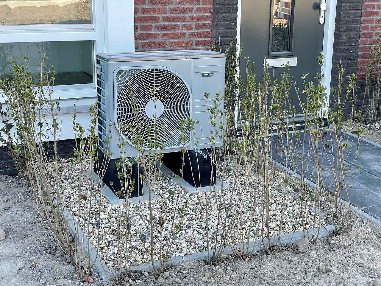 Unité extérieure de pompe à chaleur sur plots devant une maison en brique rouge, dans un massif de gravier planté de jeunes arbustes