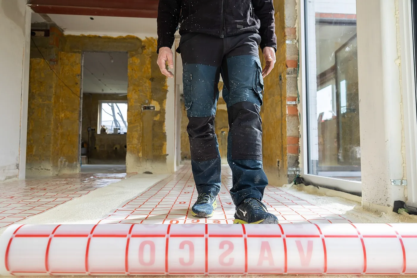 Ouvrier marchant sur l'isolant quadrillé d'un plancher chauffant qu'il déroule dans une maison en rénovation aux murs mis à nu