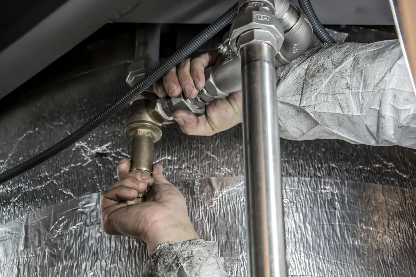 Mains d'un plombier serrant un raccord en laiton sur un tube inox, sous un plafond doublé d'isolant réfléchissant