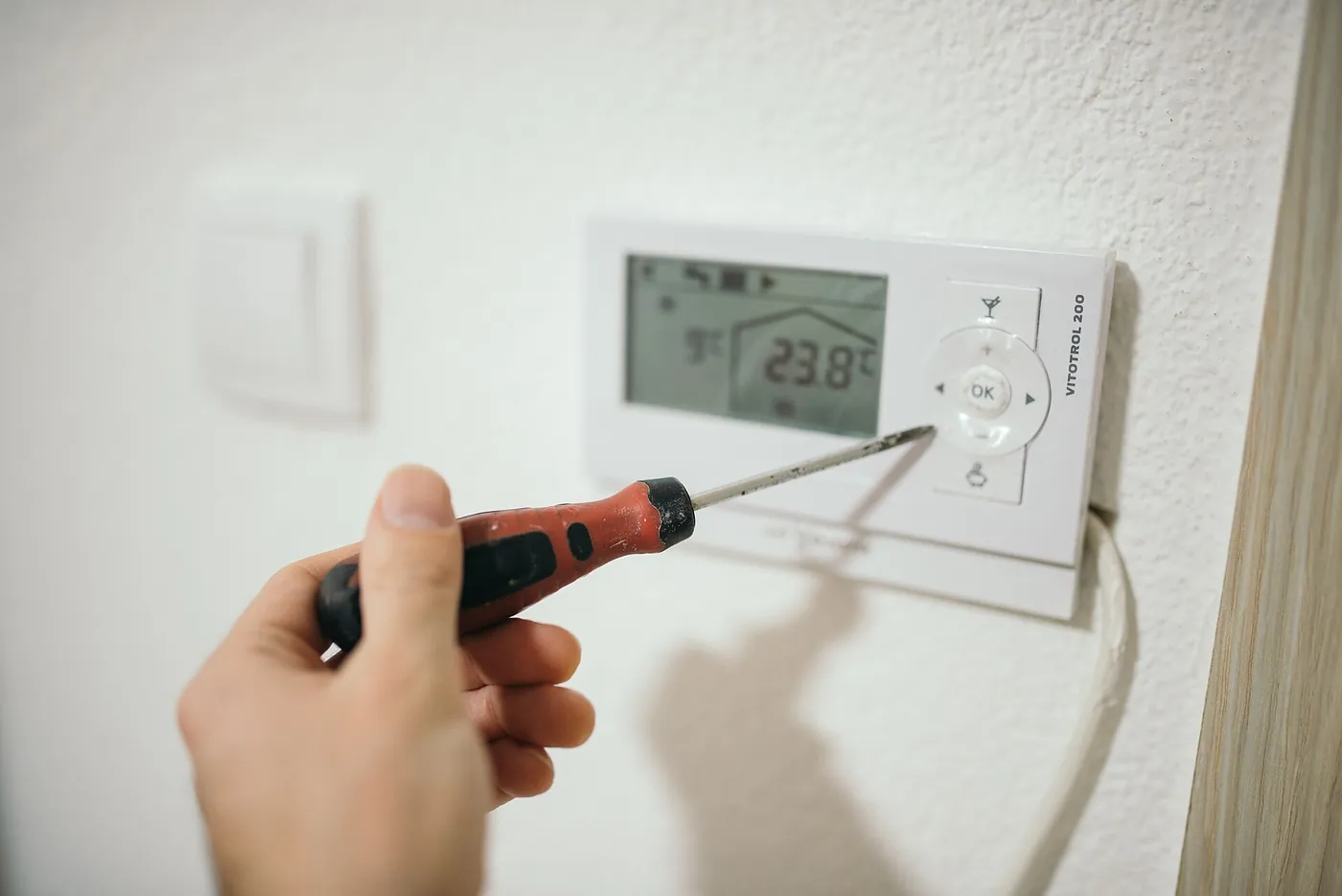 Main tenant un tournevis à manche rouge devant un thermostat d'ambiance mural blanc dont l'écran affiche 23,8 °C