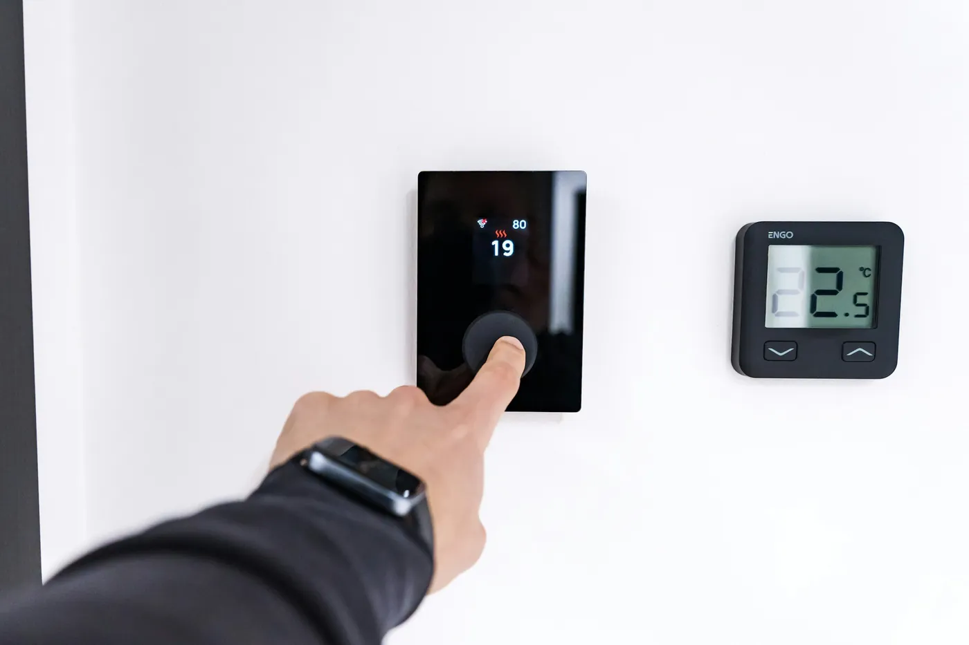Main réglant un thermostat tactile noir sur 19 °C, à côté d'un second thermostat à écran affichant 22,5 °C, sur un mur blanc