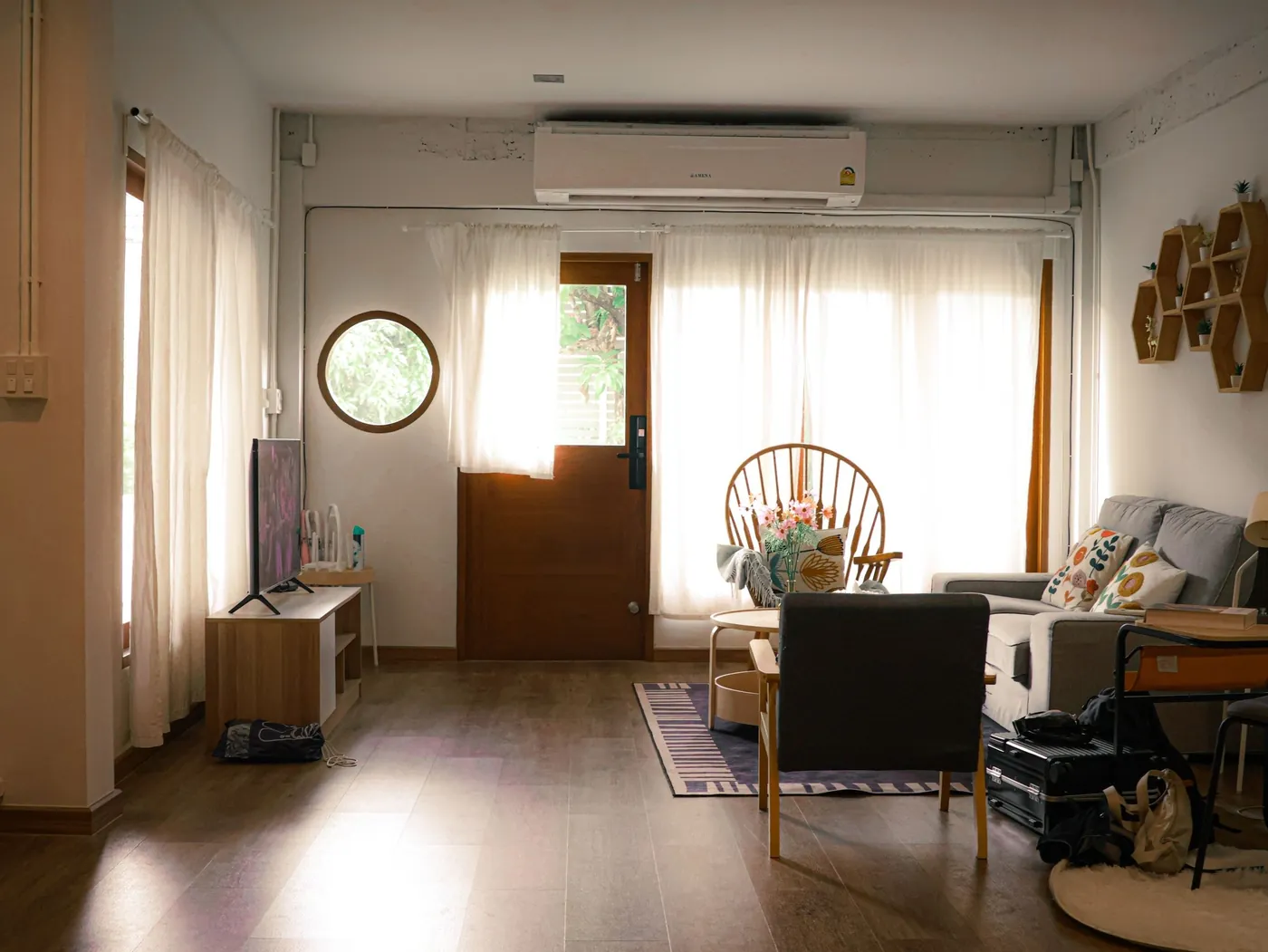 Séjour au parquet sombre, voilages blancs, hublot rond et porte vitrée, unité intérieure murale au-dessus, fauteuil en rotin et canapé gris