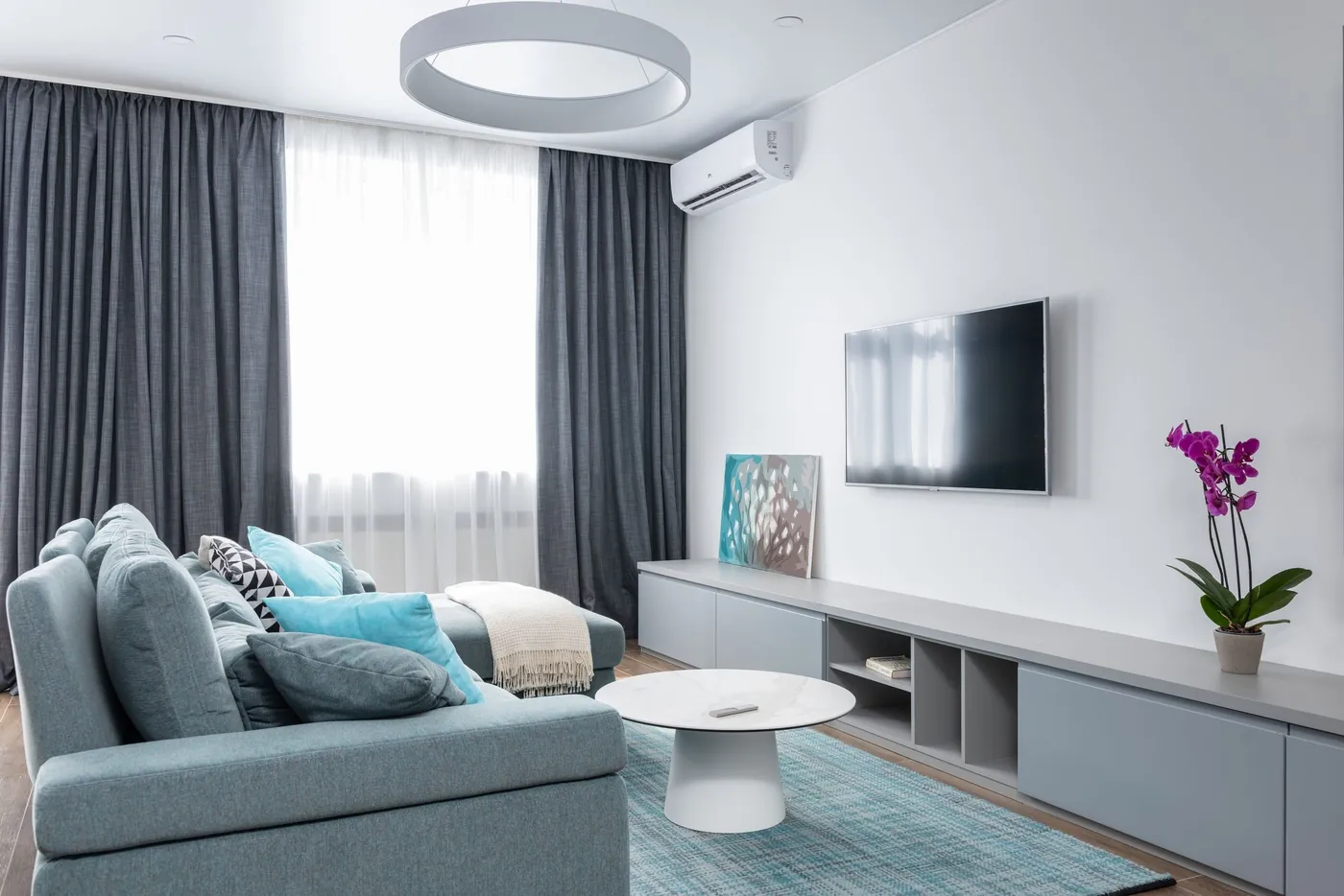 Unité intérieure murale blanche au-dessus d'une baie à voilages et rideaux gris, salon clair avec canapé turquoise et télévision