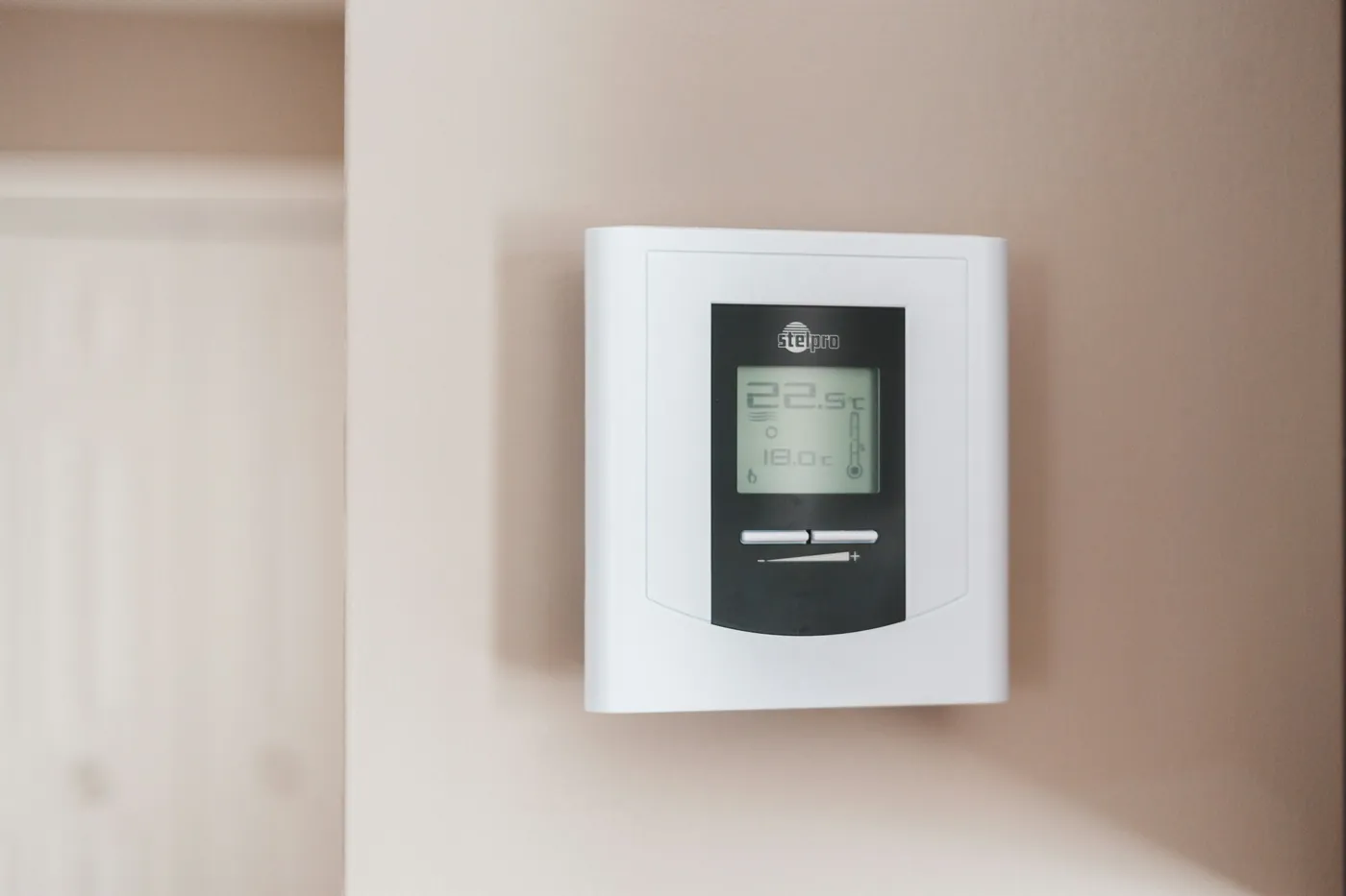 Thermostat numérique blanc à écran gris fixé sur un mur rose pâle, affichant 22,5 °C et 18,0 °C, porte floue à l'arrière-plan