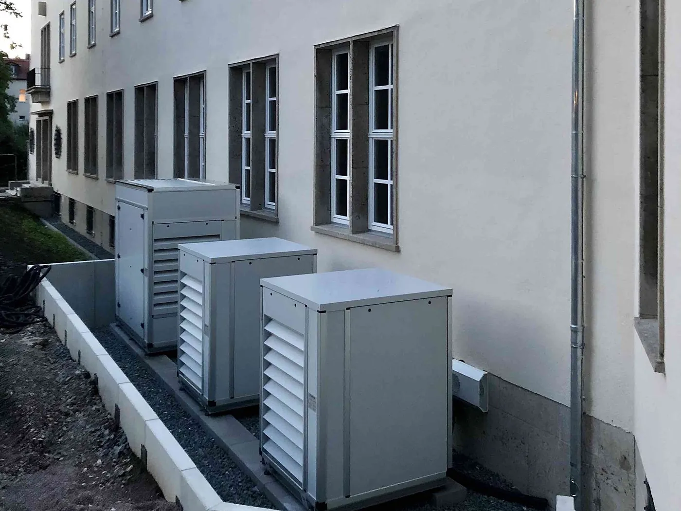 Trois unités extérieures grises à grilles sur une bande de gravier au pied d'un bâtiment ancien enduit, côté arrière