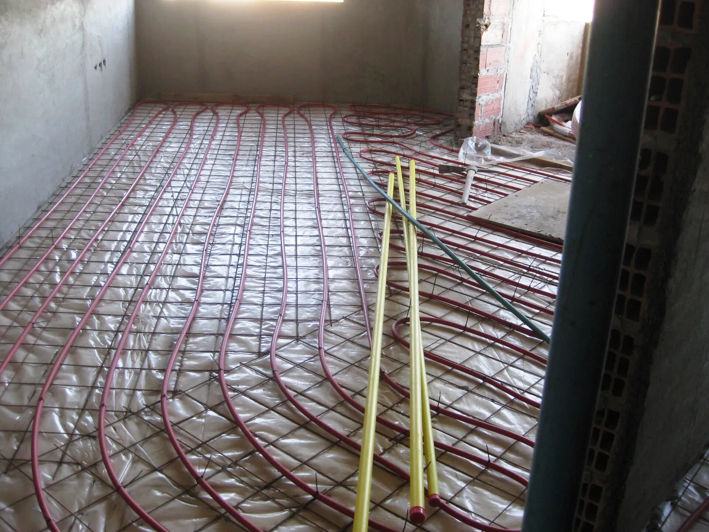Tubes rouges de plancher chauffant fixés sur un treillis métallique, dans une pièce brute aux murs de béton et de brique