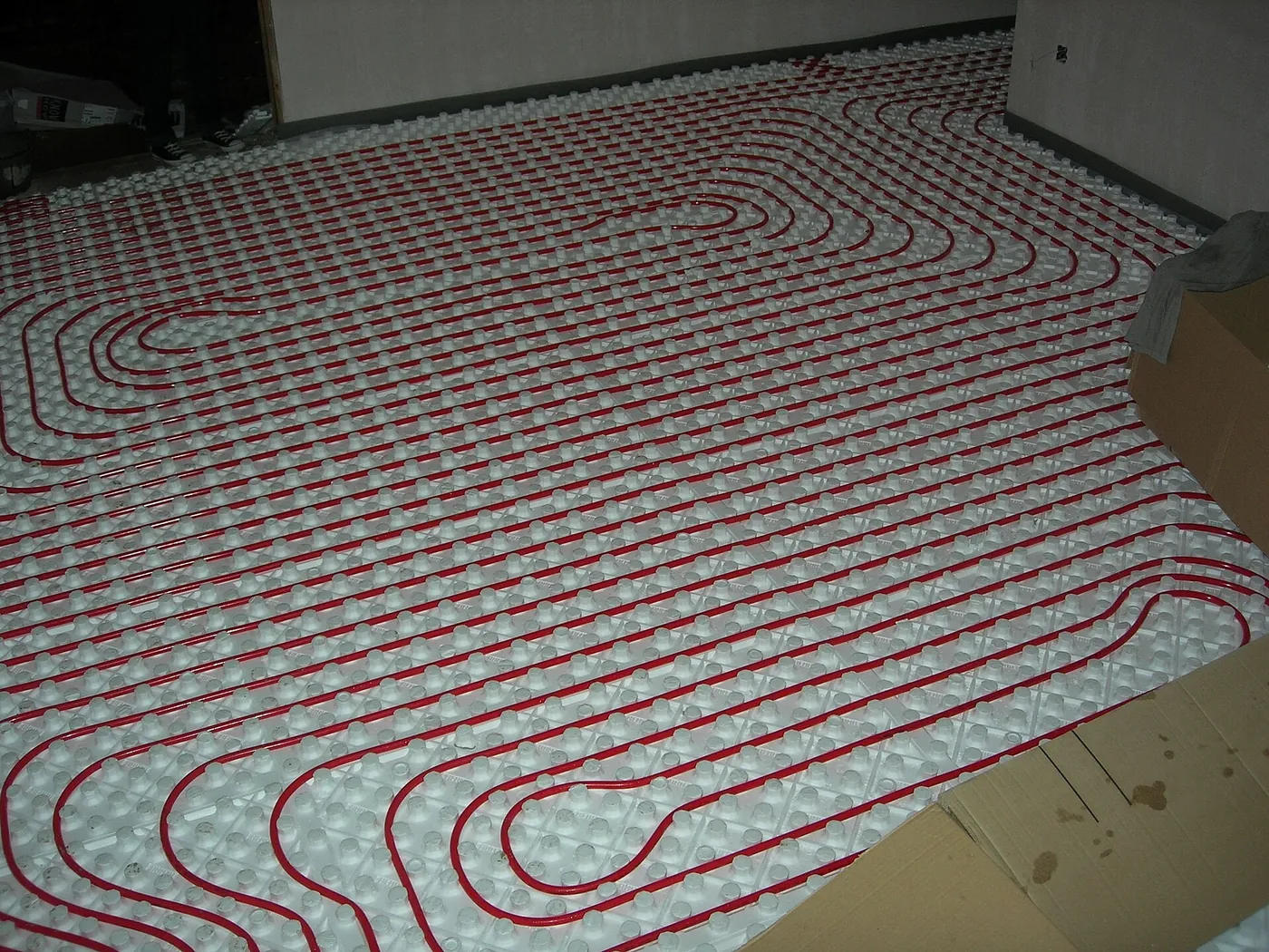 Tubes rouges de plancher chauffant posés en serpentins réguliers sur des plaques à plots blanches dans une grande pièce en travaux