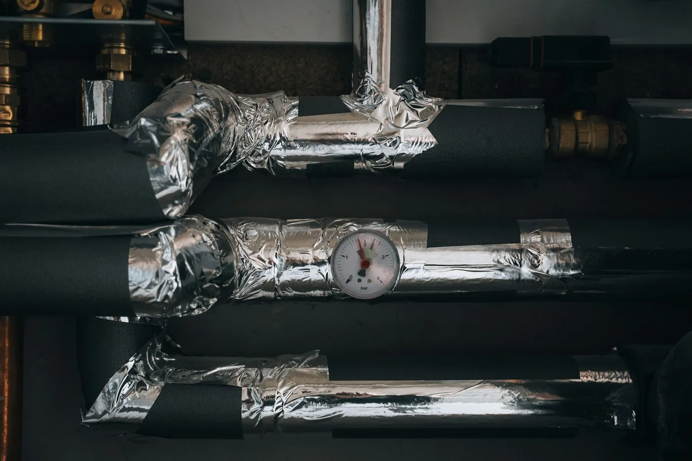 Tuyaux de chauffage gainés de mousse noire et d'aluminium, avec un manomètre à aiguille rouge, dans un local technique