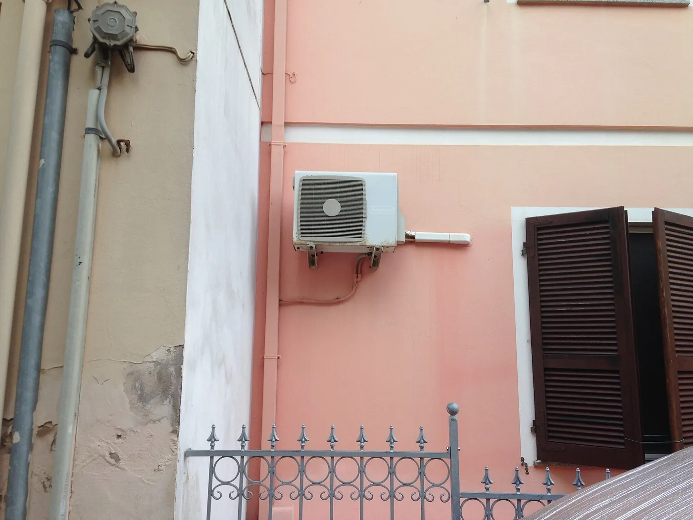 Unité extérieure sur console contre une façade rose aux volets bruns persiennés, grille en fer forgé à pointes au premier plan