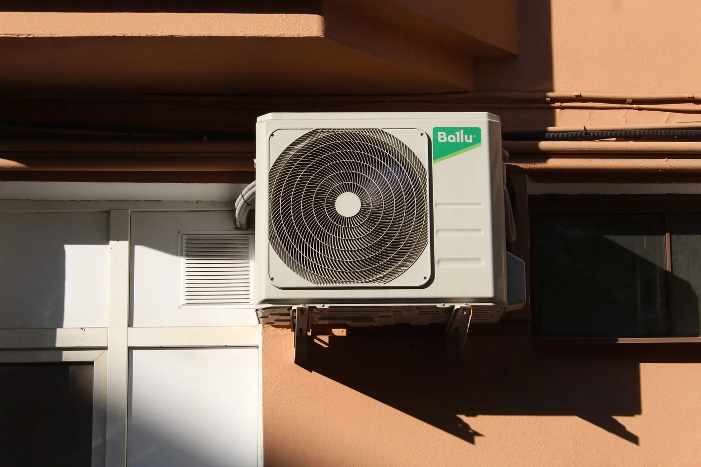 Unité extérieure de climatiseur réversible sur consoles contre une façade enduite terre cuite, tuyauteries et ombre portée en plein soleil