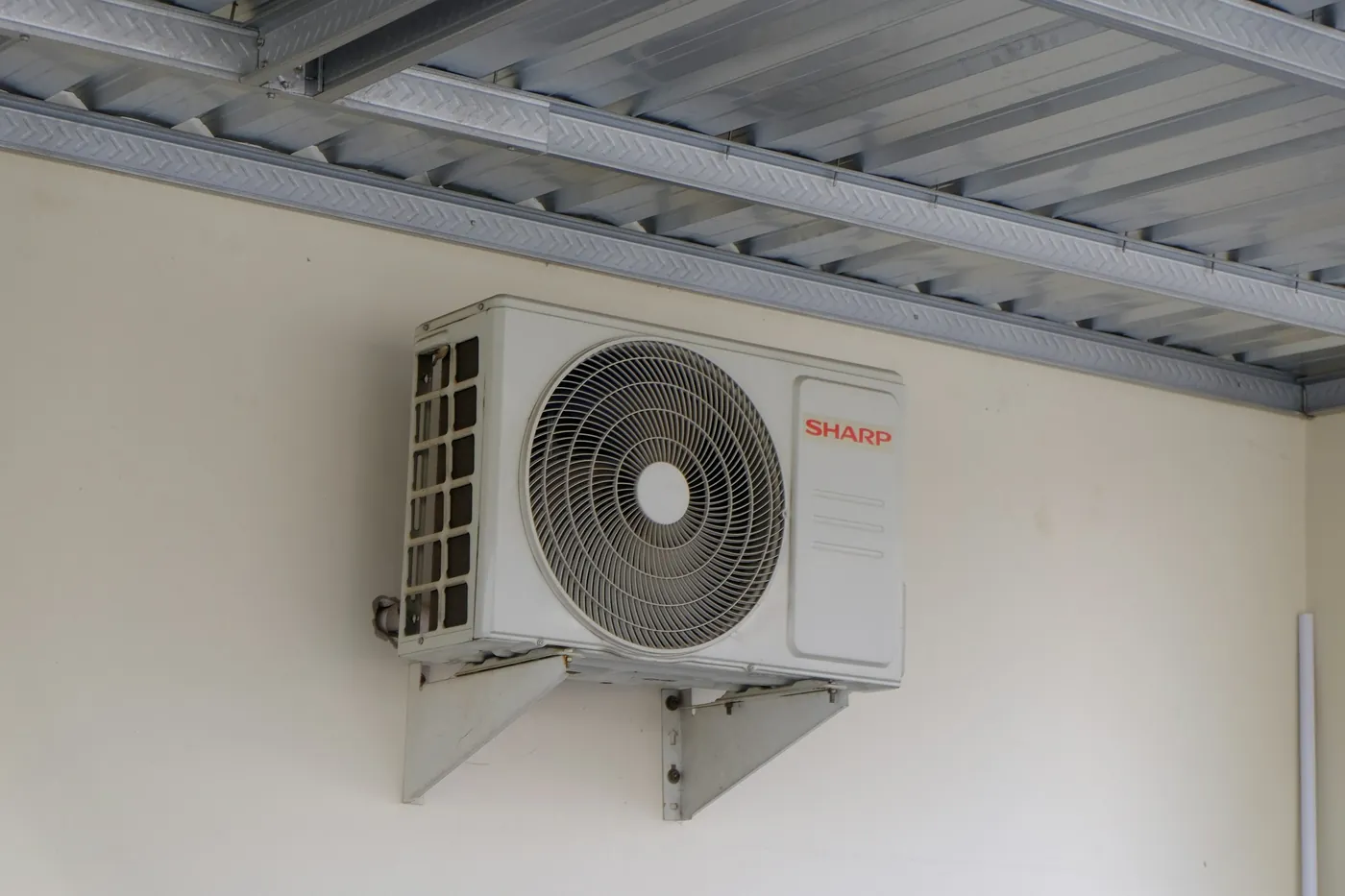 Unité extérieure de climatiseur réversible sur équerres contre un mur crème, à l'abri d'un auvent en tôle galvanisée nervurée
