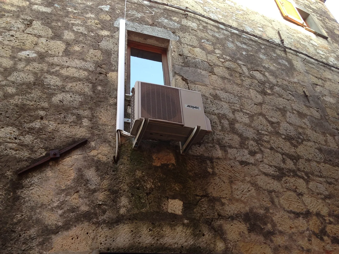 Unité extérieure beige sur équerres contre un vieux mur en moellons, sous une fenêtre étroite, goulotte blanche et ancre de tirant rouillée