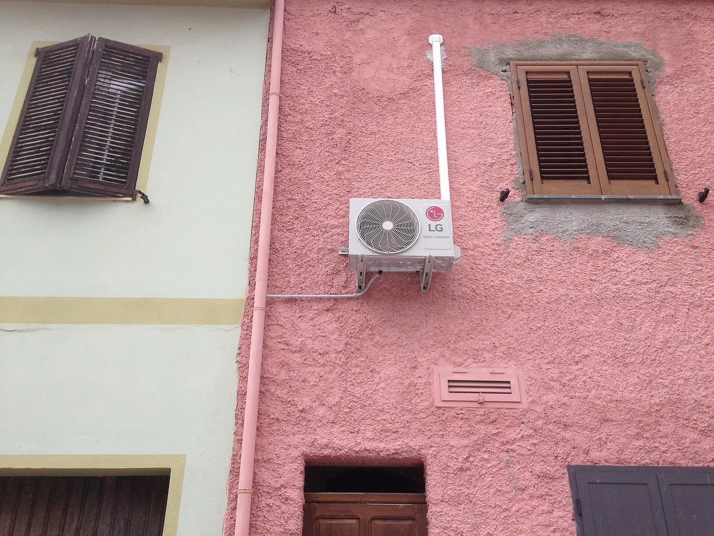 Unité extérieure sur équerres contre un mur crépi rose, goulotte blanche, volets en bois persiennés et façade voisine vert pâle