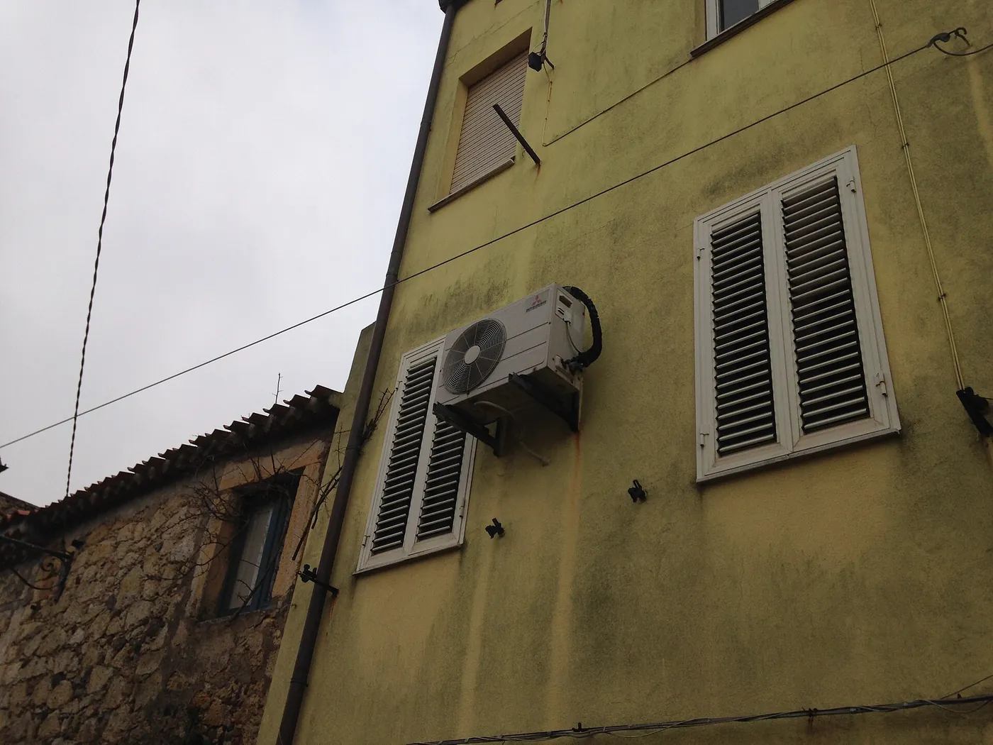 Unité extérieure sur équerres près de persiennes blanches, façade enduite vert olive tachée d'humidité, maison en pierre et ciel gris
