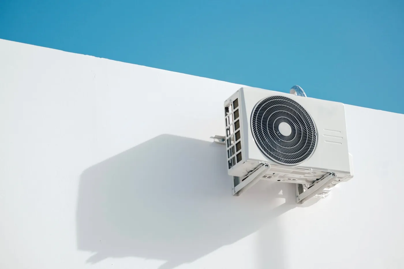 Unité extérieure blanche de pompe à chaleur fixée sur console contre un mur blanc, sous un ciel bleu