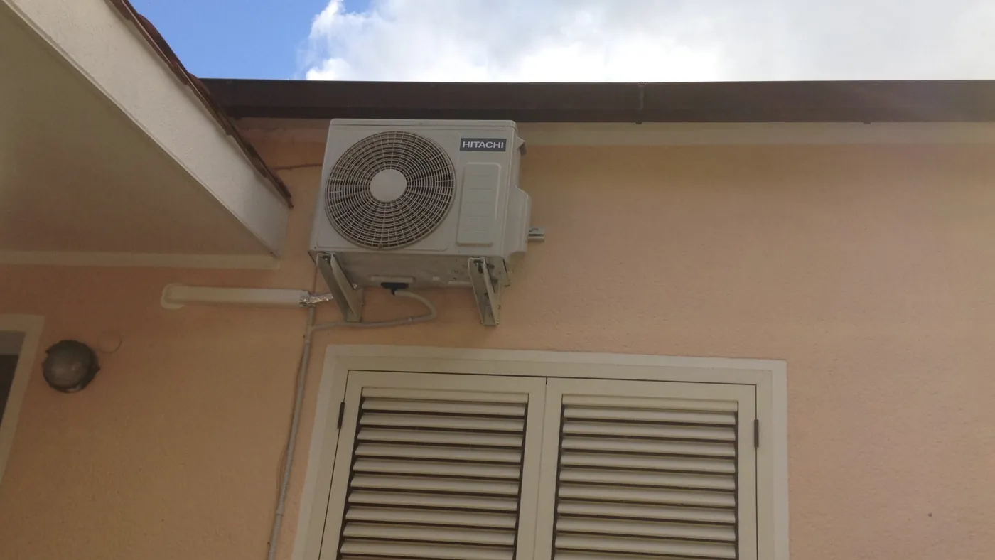 Unité extérieure sur équerres sous l'avant-toit d'une maison au crépi abricot, au-dessus d'une fenêtre à persiennes blanches